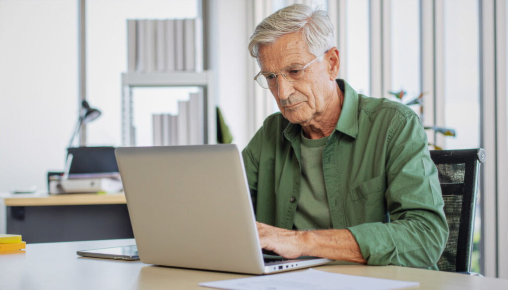 Elderly man sitting at a desk using a laptop.
